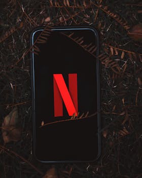 Close-up of a smartphone displaying Netflix on a leafy outdoor ground.